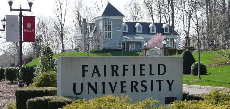 A stone sign reading "Fairfield University" stands proudly in front of a large white building with a gray roof, surrounded by lush greenery and trees. Banners displaying the university logo flutter in the background. The scene perfectly encapsulates the tradition and excellence highlighted in the Fairfield University common data set.