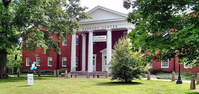 A red brick building with white columns and a sign reading "Kennedy Center" prominently showcases Hiram College's profile. A "Welcome New Students" banner hangs on the facade, while trees and greenery create a welcoming foreground.