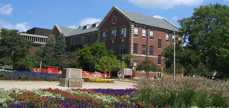 A brick academic building with large windows stands amidst a landscaped garden with various plants and flowers. A pathway leads to the entrance, and people are seen walking in the background, embodying the picturesque Illinois State University profile.