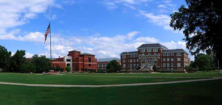 A courtyard with grass and walkways, featuring an American flag on a pole to the left and two multistory brick buildings under a blue sky dotted with clouds, reminiscent of the Mississippi State University common data set.