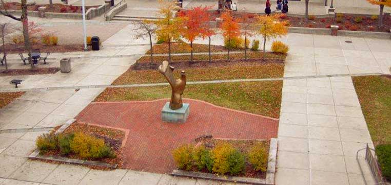 A bronze sculpture stands in the center of a brick-paved area within a plaza at Ohio Wesleyan University. Surrounding the sculpture are autumn-colored trees and neatly trimmed hedges, with scattered benches and walkways, adding to the aesthetic charm often noted in their Common Data Set.