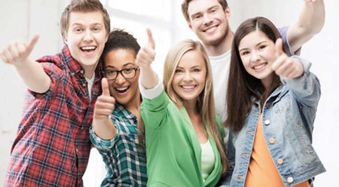 Five people standing close together, all smiling and giving thumbs up, in a brightly lit indoor setting at Saint John Fisher College.