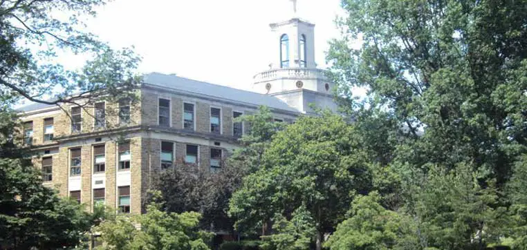 A large, three-story brick building with a clock tower is partially obscured by trees on a sunny day, presenting a serene backdrop that could easily be found within the idyllic campus of Ursinus College.