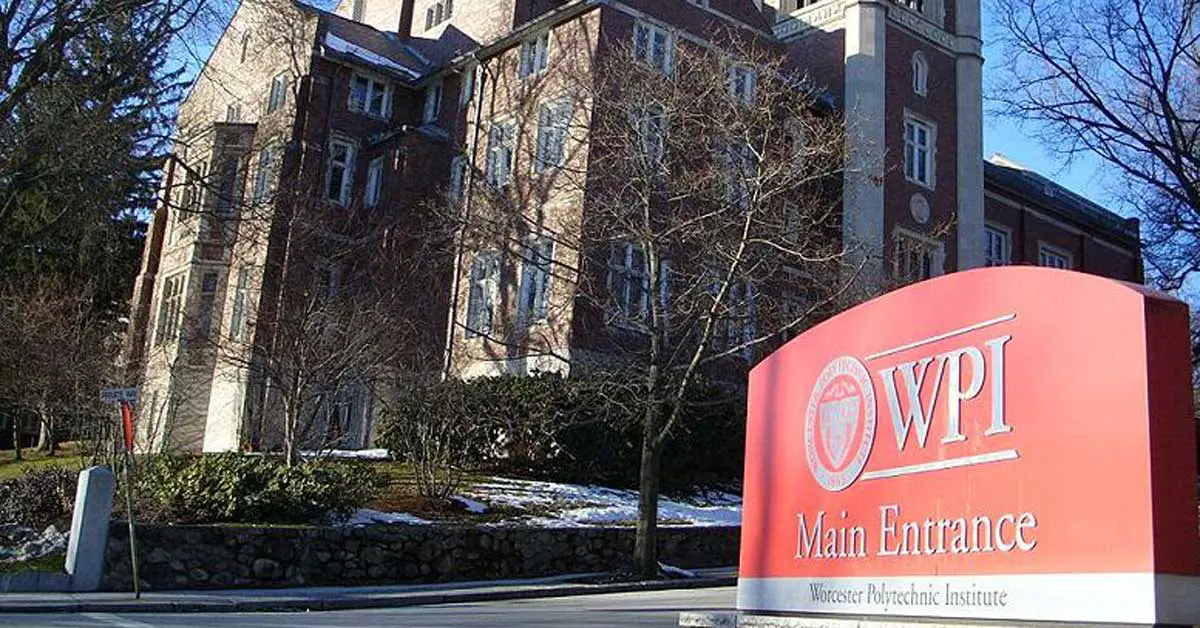 A red sign reading "WPI Main Entrance Worcester Polytechnic Institute" stands proudly in front of a large, multi-story brick building, framed by trees and patches of snow on the ground.