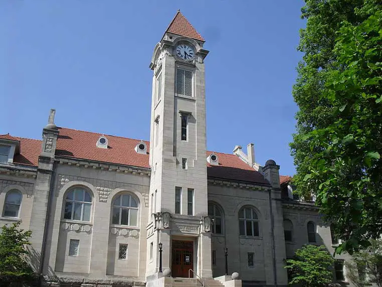 A historic building with a clock tower and red-tiled roof, surrounded by greenery, under a clear blue sky, reminiscent of the timeless architecture seen on the Indiana University campus.