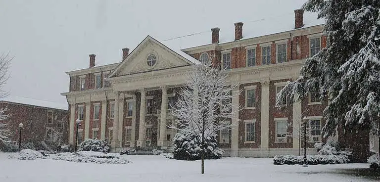 A large brick building with white columns and a triangular pediment at Roanoke College is surrounded by snow-covered trees and ground on a snowy day, offering a picturesque backdrop that could easily be featured in their common data set imagery.