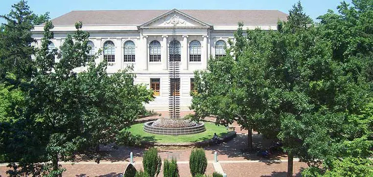 A large, neoclassical building with multiple large windows and a central entrance stands majestically. In front, a brick pathway leads to a circular fountain surrounded by green trees and neatly trimmed bushes, reminiscent of the stunning campus scenery at the University of Arkansas.