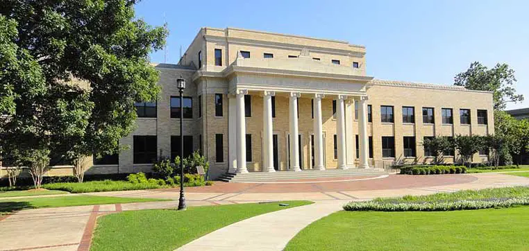 A three-story building with large columns at the entrance, surrounded by a well-maintained lawn and paths, under a clear blue sky, reflects the picturesque charm of Austin College.