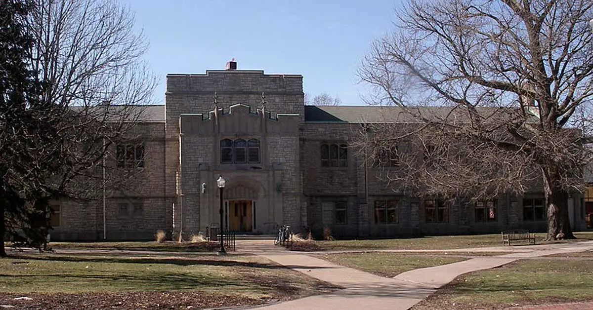 A stone building with gothic architectural elements, featuring a central entrance flanked by two trees and intersecting pathways leading to the door. The surrounding area has a few bare trees, much like the serene campus of Knox College described in their common data set.