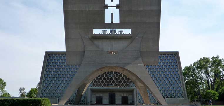 Concrete facade of a modernist building with hexagonal patterned walls and an arch above the entrance, framed by trees on either side, reminiscent of the architectural style found at Saint John's University.