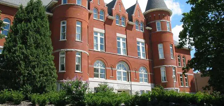 A historic red-brick building with multiple arched windows and a conical tower, surrounded by greenery, reminiscent of the iconic architecture found at Washington State University.