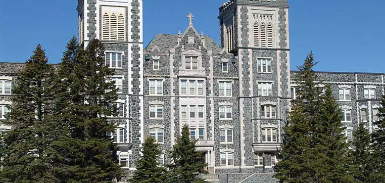 A large stone building with two tall tower-like structures stands against a clear blue sky, surrounded by evergreen trees at the College of St. Scholastica.