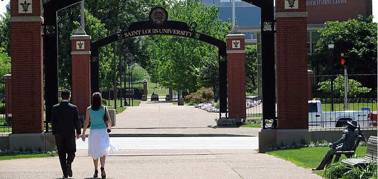 A man and woman hold hands as they stroll towards the entrance arch of Saint Louis University, framed by green trees and a paved walkway. The scene reflects the welcoming atmosphere often highlighted in the Saint Louis University Common Data Set.