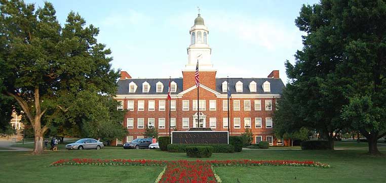 A large brick building with a white cupola and flagpole stands proudly, surrounded by trees and cars parked in front. In the foreground, manicured lawns and flower beds form decorative patterns, reflecting the charm you might find at Transylvania University.