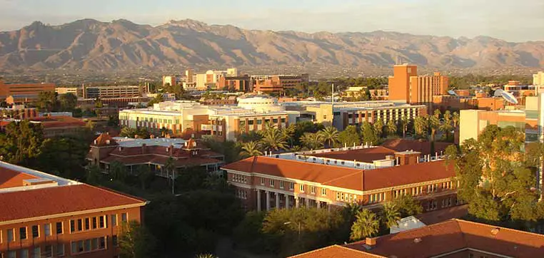 Aerial view of the University of Arizona campus with red-roofed buildings, surrounded by greenery and set against a backdrop of mountains under a partly cloudy sky.