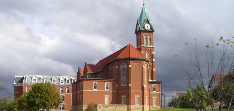 A red brick building with a clock tower and steep green roof stands under a cloudy sky at Loras College, surrounded by trees and grass.