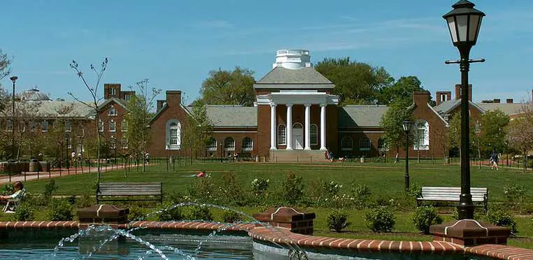 A brick building with white columns stands behind a lawn and benches, framed by trees and a fountain in the foreground on a sunny day, embodying the picturesque campus often seen on a University of Delaware profile.