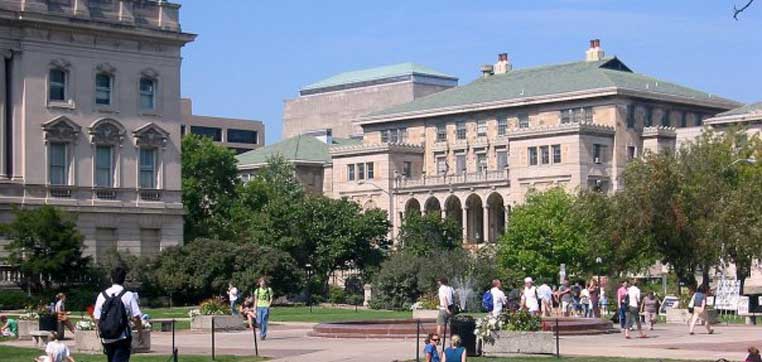 A photo of the University of Wisconsin-Madison campus with historic buildings, a green lawn, trees, and people walking and sitting.