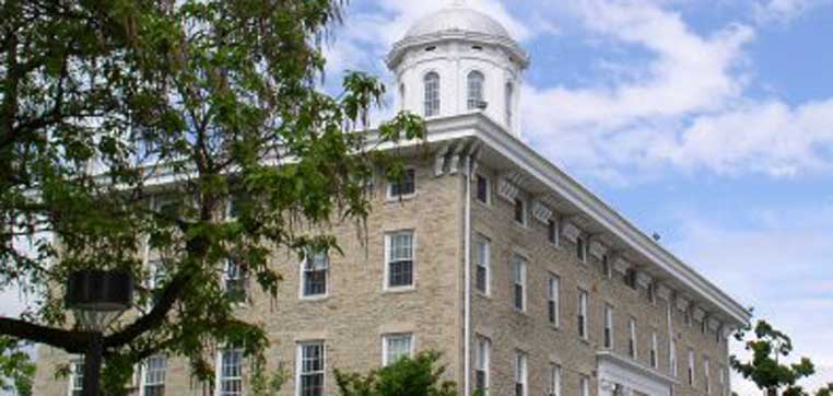 A stone building with multiple windows and a white cupola on the roof stands elegantly amidst lush trees, under a partly cloudy sky, epitomizing the charm of Lawrence University's campus.