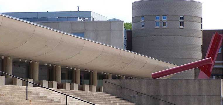 Modern building at SUNY at Fredonia featuring a curved concrete structure, cylindrical tower, and red abstract sculpture, with steps leading up to the entrance.