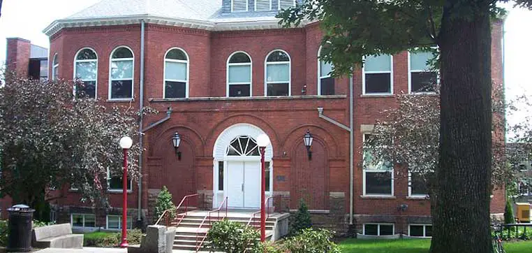 A two-story brick building with arched windows and a central white door, surrounded by trees and landscaping, could easily be part of the charming campus at Indiana University of Pennsylvania.