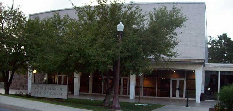 Exterior view of the John F. Kennedy Student Center at Saint Francis University, featuring a large building with a sign, lush trees, and a classic lamppost in front.