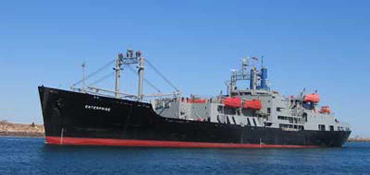 A large, gray cargo ship with cranes on its deck sails in calm blue waters under a clear sky, a scene reminiscent of the training vessels used by the Massachusetts Maritime Academy.