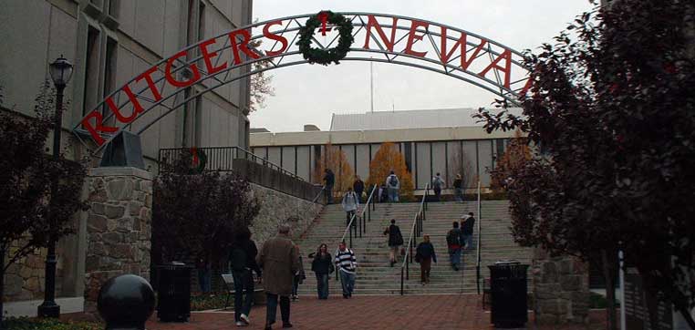 A group of people walk up stairs under an archway that reads "Rutgers University-Newark." The scene is outdoors with trees on either side, capturing the vibrant campus life detailed in the Rutgers University-Newark Common Data Set.