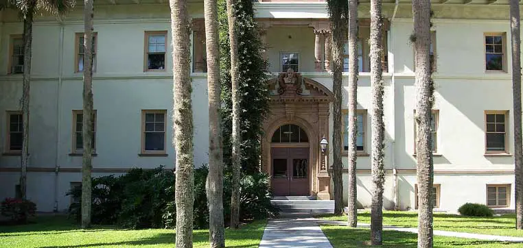 A light-colored, multi-story building with a decorative arched entrance surrounded by tall, slender palm trees and green lawn, reminiscent of the picturesque architecture found on the campus of Stetson University.