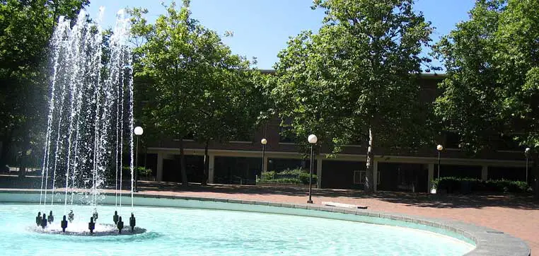 A circular fountain with water jets shooting upwards in front of a Western Washington University building, surrounded by trees on a sunny day.