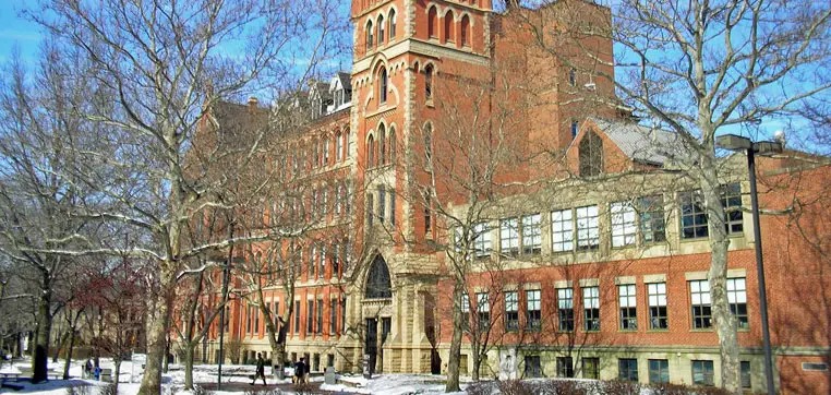 Red-brick building with arched entrance and tall central tower, surrounded by bare trees under a clear blue sky; snow covers the ground. This picturesque scene resembles the iconic architecture of John Carroll University, known for its competitive acceptance rate.