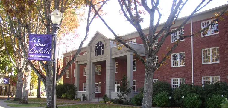 A brick building with white columns and large windows on a sunny day. A blue banner on a lamppost reads, "It's your Linfield University. Welcome Home." Trees with autumn foliage are in the foreground.