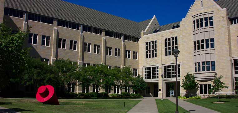 A large beige and gray stone building with tall windows is surrounded by greenery and a sidewalk. A red sculpture is placed on the lawn in front of the University of St. Thomas building.