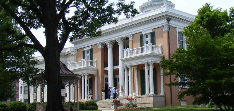 A large, white-columned mansion with a beige exterior, surrounded by trees and greenery, sits majestically on the grounds of Belmont University. Several people are gathered on the front steps under a clear sky.