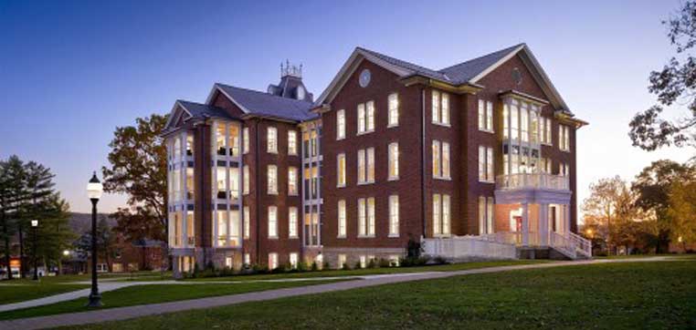 A large, modern brick building with many windows is illuminated at dusk on the Juniata College campus. It has multiple stories and a front porch. A well-maintained lawn and a streetlamp are in the foreground.