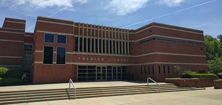 The image shows the exterior of Trexler Library, a red-brick building with large windows and a set of stairs leading to the entrance under a clear, sunny sky, capturing the charming architectural profile of DeSales University.