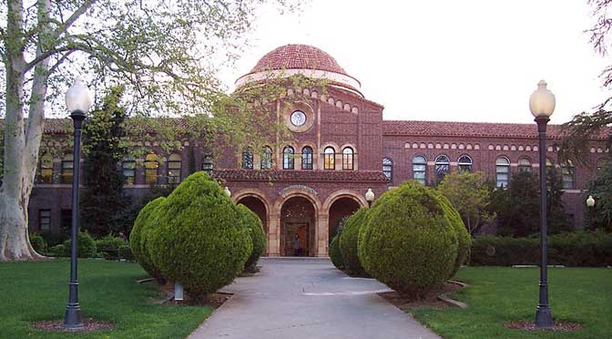 A red-brick building with a domed roof and clock above the entrance, featured in the California State University-Chico profile, is flanked by trees and bushes. The path leading to the entrance is lined with street lamps and manicured shrubs.