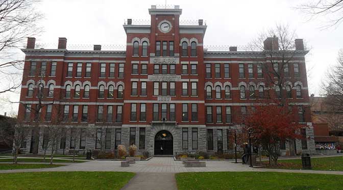 A large, red-brick building with a clock tower stands behind a grassy area and a path leading to the entrance. The building is symmetrical with multiple windows and arched entryways. Trees surround the area, creating a picturesque scene that perfectly complements Clark University’s profile.