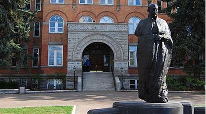 A statue stands in front of the entrance to a brick building labeled "Gonzaga College," featuring arched windows and a staircase leading up to the door, perfectly capturing the Gonzaga University profile.