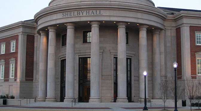 A neoclassical building with large columns at the entrance and the name "Shelby Hall" above them, showcasing the distinguished profile of the University of Alabama. Two lamp posts are visible in the front.