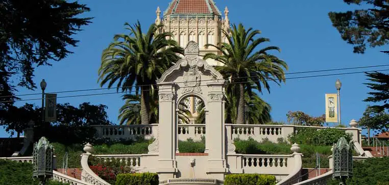 A grand staircase with manicured hedges and ornate sculptures leads up to a domed building, surrounded by palm trees and greenery, under a clear blue sky—a picturesque scene reminiscent of the University of San Francisco's profile.