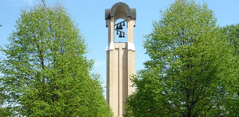 A tall bell tower made of brick, reminiscent of the iconic structures at Concordia College, stands gracefully with multiple bells inside. It is surrounded by green trees and a clear sky, creating a picturesque profile.