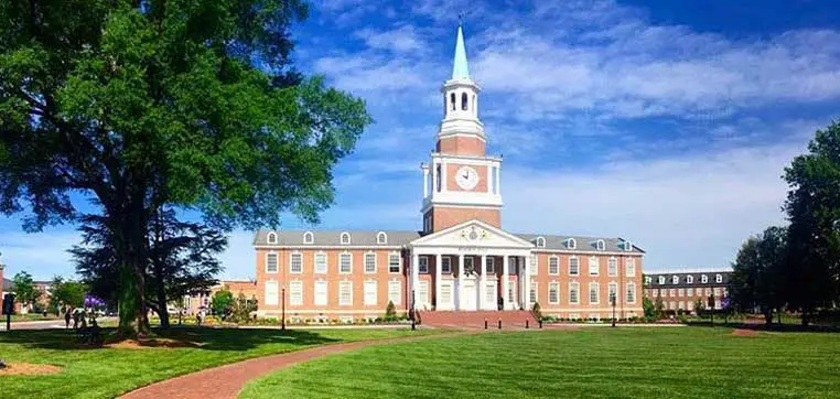 A red-brick building with white columns and a tall clock tower stands on a well-manicured lawn at High Point University, set against a bright blue sky with some trees on either side.