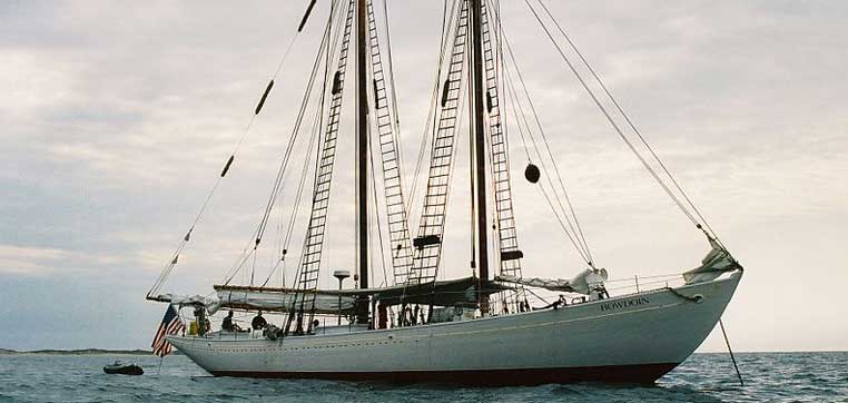 A large sailing ship with two tall masts and intricate rigging, reflecting the esteemed tradition of the Maine Maritime Academy, is anchored in open water under a cloudy sky.