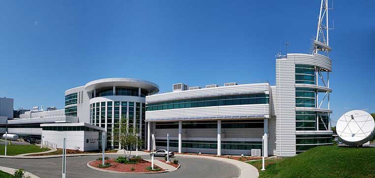 A modern multi-level building with glass windows and a white exterior stands proudly under the clear blue sky, adjacent to a large white satellite dish. A car is parked near the entrance, capturing the essence of innovation at University at Albany.