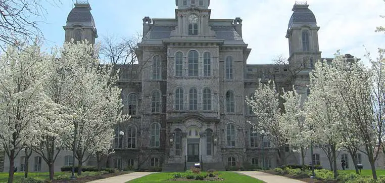 A large, historic stone building with arched windows stands majestically on the Syracuse University campus, surrounded by blooming white trees and green lawns under a clear sky.