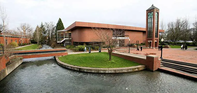 A modern brick building with a large clock tower sits alongside a gently flowing stream at Willamette University, featuring a landscaped area and a few people walking nearby.