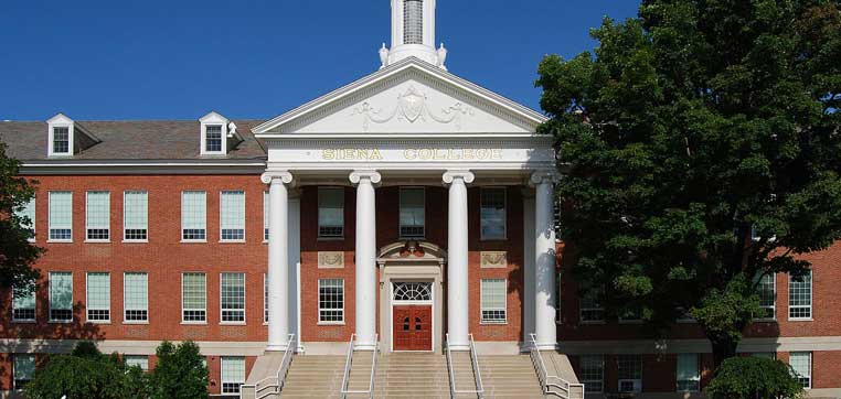 A red-brick institutional building with a white-columned entrance proudly displays "Siena College" above the door. The building is surrounded by green trees, and the sky is clear, capturing an idyllic moment at Siena College.