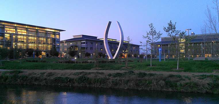 A modern university campus at dusk features contemporary buildings with large windows and a curved metal sculpture in the center, with a river in the foreground. The scene echoes the forward-thinking spirit of the University of California-Merced.