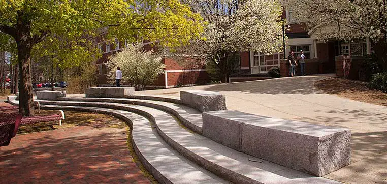 Outdoor stone seating area with curved steps on a paved pathway, surrounded by trees and brick buildings in the background with a few people walking, much like the tranquil spots you’d find at Western New England University.